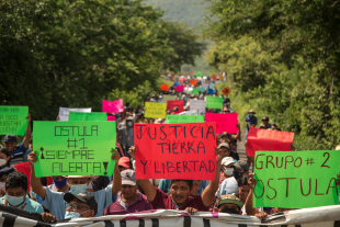 Auf dem Foto sind Demonstrant*innen zu sehen, die rote, grüne und weitere bunte Demo-Schilder mit beiden Händen in die Höhe halten. Im Hintergrund und weiteren Straßenverlauf sind viele weitere Demonstrant*innen zu sehen. Die Straße ist von grünen Bäumen umgeben.