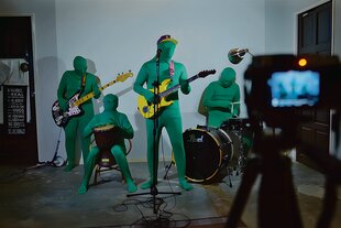 Vier Musiker*innen in grünen Ganzkörper-Anzügen spielen vor einer weißen Wand und werden dabei gefilmt.