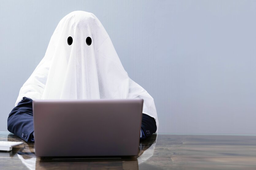 Ein Mensch, der sich mit einem weißen Laken über dem Kopf als Gespenst verkleidet hat, sitzt hinter einem Laptop