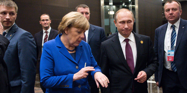 Angela Merkel und Wladimir Putin laufen einen Gang entlang, neben ihnen weitere Personen
