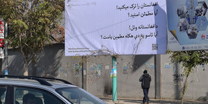 Ein Plakat mit der Aufschrift „Sie verlassen Afghanistan? Sind sie sicher?“. Davor Autos auf der Straße