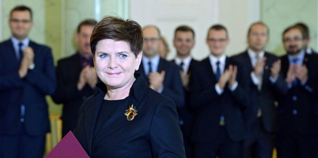 Beata Szydło schaut in die Kamera, hinter ihr stehen Politiker und Politikerinnen und klatschen für sie.