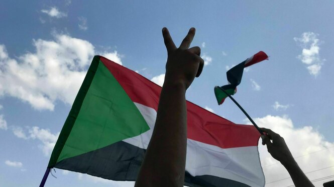 Eine Person zeigt das "Viktory-Zeichen" mit den Fingern, eine sudanesische Flagge weht im Hintergrund