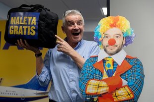 Michael O'Leary hält ein Gepäckstück in den Händen, grimassiert und steht neben einer Clownsfigur aus Pappe