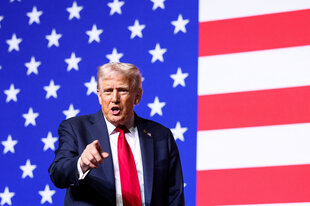 Donald Trump steht vor einer riesigen US-Fahne und gestikuliert