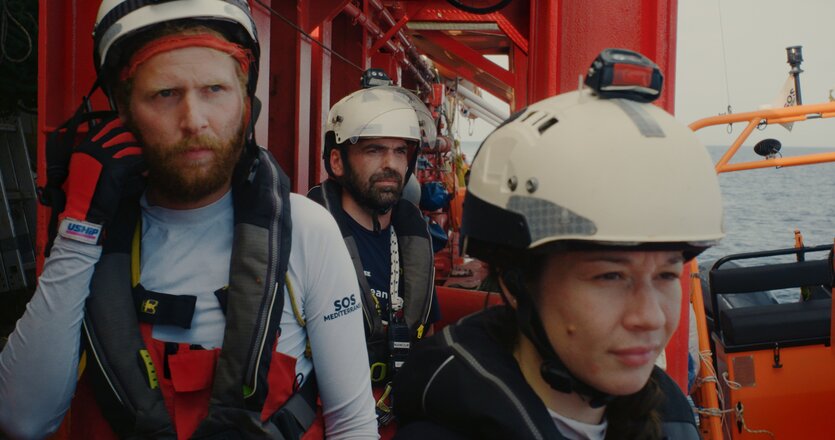 Menschen mit angespannten Mienen auf einem Schiff. Sie tragen Helme und schauen aufs Meer.