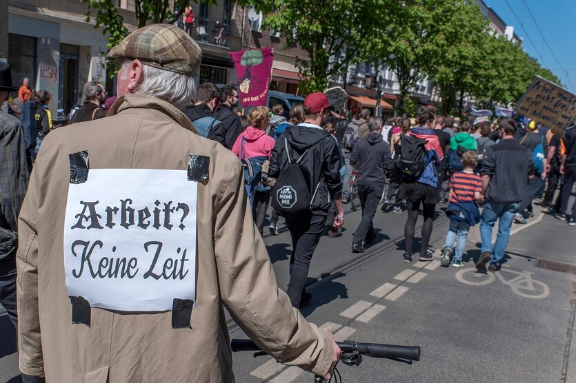 Ein Mann steht am Rande eines Demonstrationszugs, auf seinem Rücken ist ein Zettel gehftet, darauf steht: "Arbeit-kein Zeit"