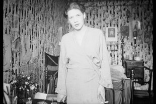 Eine Schwarz-weiß-Fotografie von Mopsa Sternheim 1933 in einer Wohnung in Paris. Sternheim trägt kurze Haare, eine Art Wickelkleid oder Bademantel, in der linken Hand eine Zigarette; ihr Kopf ist leicht geneigt, sie schaut etwas schräg in die Kamera ihrer