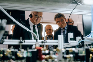 Im Vordergrund zu sehen sind technische Apparaturen eines Labors des Centre Européen des Sciences Quantiques in Straßburg, im Hintergrund eine Personengruppe, zu der auch Baden-Württembergs Ministerpräsident Winfried Kretschmann gehört, die die Apparaturen betrachtet.