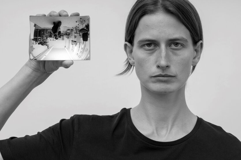 Schwarzweißes Foto, eine Frau enface, sie hält einen Spiegel in der Hand, in dem der Raum Kopf steht