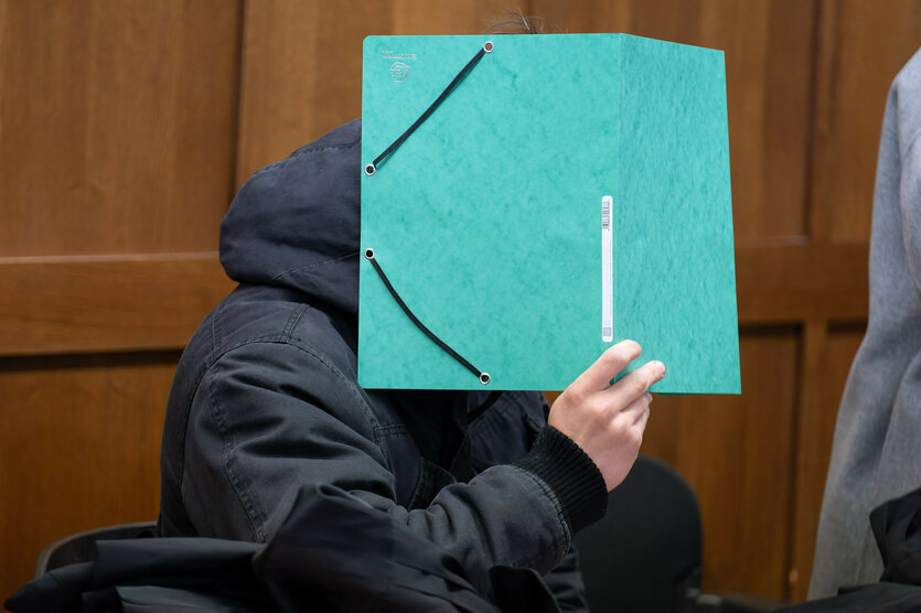 Ein Mann in einer schwarzen Jacke hält sich einen grüne Mappe vor das Gesicht