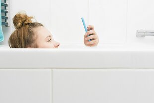 Eine junge Frau liegt in der Badewanne und schaut auf ihr Smartphone, zu sehen ist die Hälfte ihres Gesichts und die Hand, die über den Beckenrand ragt