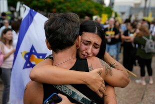 Eine junge Frau umarmt einen Mann, sie stehen auf einem Platz in Tel Aviv und eine unscharfe israelische Flagge ist zu sehen