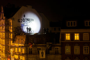 Das Bild zeigt nachts ein Gebäude mit Graffiti, darunter die Aufschrift "G20 ANBREIFEN". Auf dem Dach stehen Polizisten, beleuchtet von einem starken Lichtstrahl. Das Gebäude ist mit Gerüst umgeben, und im Hintergrund sind erleuchtete Fenster eines weiteren Hauses sichtbar.