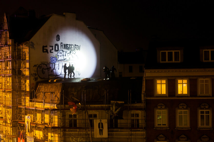 Das Bild zeigt nachts ein Gebäude mit Graffiti, darunter die Aufschrift "G20 ANBREIFEN". Auf dem Dach stehen Polizisten, beleuchtet von einem starken Lichtstrahl. Das Gebäude ist mit Gerüst umgeben, und im Hintergrund sind erleuchtete Fenster eines weiteren Hauses sichtbar.