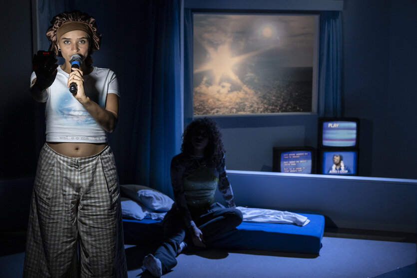 Eine Person steht mit einem Mikrofon und einem Handy in der Hand in einem dunklen Raum, während eine zweite Person auf einem Bett sitzt. Im Hintergrund ist ein Fenster mit einem leuchtenden Himmel und mehrere alte Fernseher mit dem Wort "PLAY" sichtbar. Die Szene ist in blaues Licht getaucht.