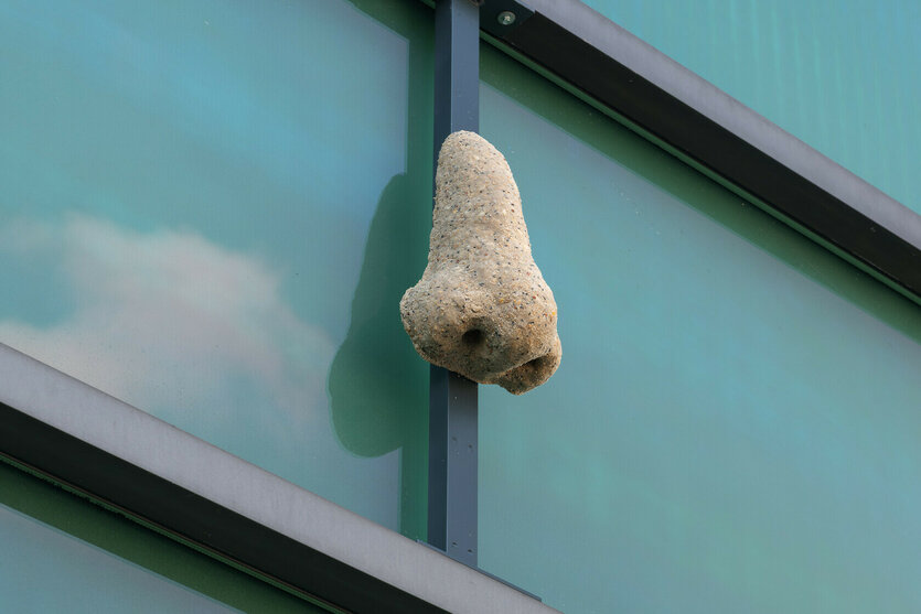 Das Bild zeigt eine große, realistische Nase aus Stein oder Beton, die an einem Metallrahmen vor einer gläsernen Fassade mit blau-grünem Reflex hängt.