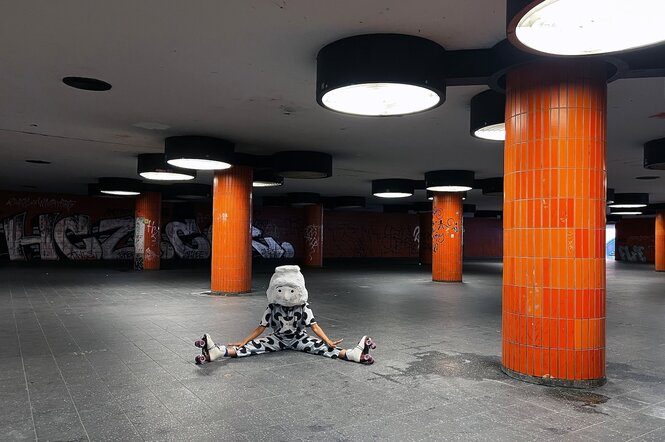 Die Comiczeichnerin Aisha Franz situt mit ausgestreckten Beinen auf dem Boden einer S-Bahnstation mit Rollenschuhen und einer weißen. Sie trägt ein weißes Outfit mit großen schwarzen Kreisen. Auf dem Kopf trägt sie das Gesicht einer ihrer schwarz-weißen Comicfiguren als überdimensional große Maske.