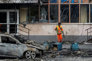 Ein Mann in orangefarbener Arbeitskleidung steht vor einem zerstörten Haus