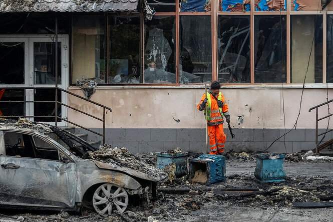 Ein Mann in orangefarbener Arbeitskleidung steht vor einem zerstörten Haus