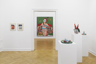 In einem Ausstellungraum stehen drei glasierte Karamikskulpturen auf weißen Sockeln. Die erste Skulptur zeigt eine blaue Figur zwischen Erdbeeren. Die zweite eine Vase, auf der Waden mit Schachbrettmuster in roten Stöckelschuhen abgebildet sind. Im Hintergrund ist ein zweiter Raum zu sehen, in dem ein Gemälde an der Wand hängt, das eine Frau zeigt, die Unterwäsche und einen roten Morgenmantel trägt. Das Innenfutter des Morgenmantels hat ein buntes Karomuster. Die braunen Haare der Frau verlaufen in einen Blauton und wickeln sich um ihren Körper, in der Vase steht eine rote Blüte.