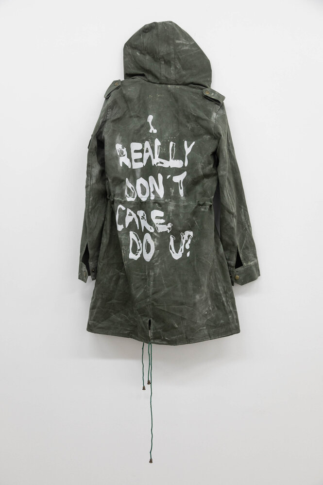 An einer Ausstellungswand hängt ein grau-grüner Parker mit Kapuze. Auf dem Rücken des Parkers sind in Weiß die Worte "I really don't care. Do you?" aufgesprüht.