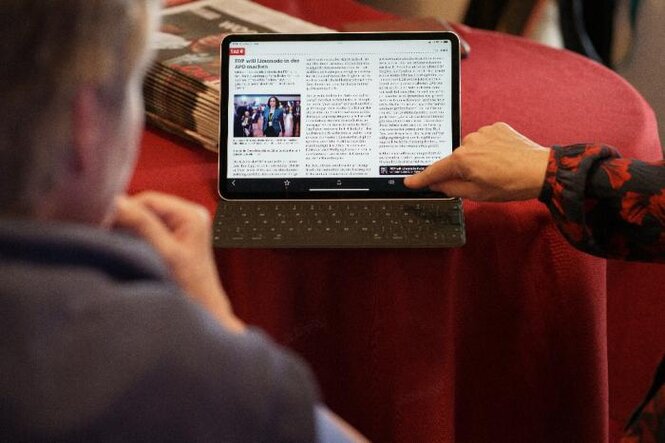 Ein Tablet mit taz App steht auf einem Tisch mit rotem Tischtuch, eine Hand bedient den Touc-Screen, eine Person schaut zu.