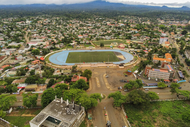 Luftaufnahme des Nationalstadions von São Tomé und Príncipe, umgeben von Wohnhäusern und Grünflächen. Die blaue Laufbahn umrundet ein Fußballfeld in der Stadionmitte. Im Vordergrund sind Verwaltungsgebäude und geparkte Fahrzeuge zu sehen, im Hintergrund erstrecken sich bewaldete Hügel unter einer bewölkten Himmelsdecke.
