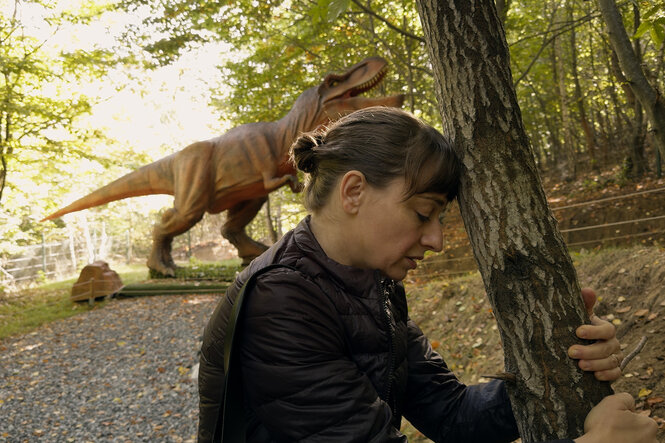 Eine Frau lehnt ihre Stirn an einen Baum, sie sieht erschöpft aus. Hinter ihr steht ein künstlicher Dinosaurier