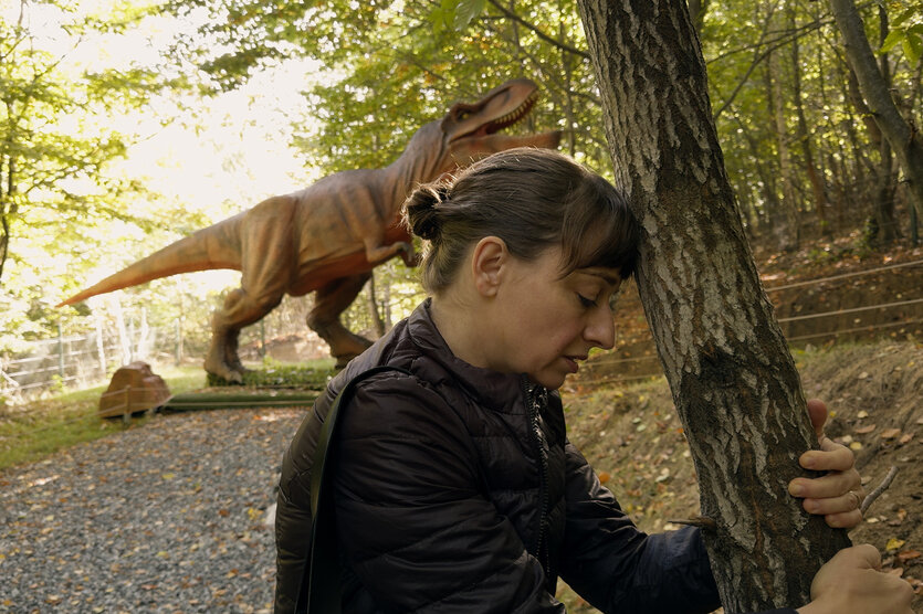 Eine Frau lehnt ihre Stirn an einen Baum, sie sieht erschöpft aus. Hinter ihr steht ein künstlicher Dinosaurier