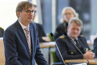 Soenke Neitzel steht im Anzug vor einem Rednerpult und spricht in ein Mikrofon, hinter ihm sitzende Leute, die den Kopf nach ihm gewandt zuhören.