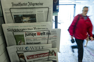 Ein Zeitungsständer mit diversen Tageszeitungen, darunter die tageszeitung taz. Neben dem Zeitungsständer läuft eine Person in roter Jacke vorbei.