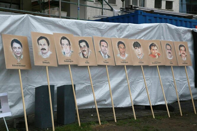 Plakate an Stangen mit Porträts der Opfer des NSU lehnen an einem verhüllten Bauzaun