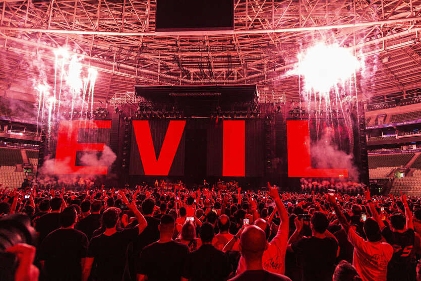 Eine Konzertbühne in rotes Licht getaucht, mit dem Schriftzug "EVIL". Im Vordergrund Fans, die ihre Arme heben