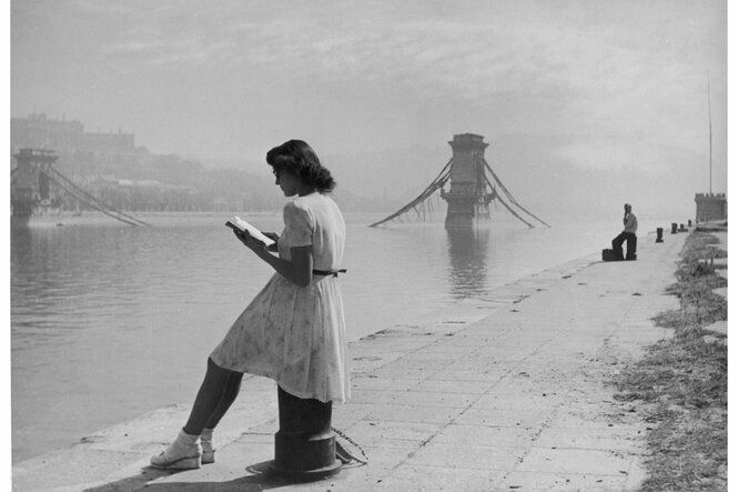 Ein Schwarzweißbild. Eine Frau sitzt an einem Fluss und liest in einem Buch.