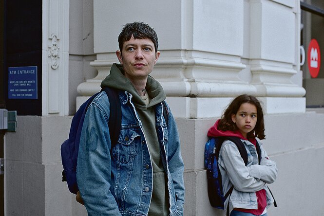 In einer Filmszene steht Cass neben Arie vor einer Hausfassade. Arie schaut Cass verunsichert mit verschränkten Armen an.