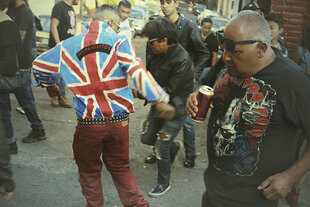 Eine Gruppe Punks tanzt auf einer Straße in Mexiko City. Ein Mann, der in der Mitte des Filmstills mit dem Rücken zur Kamera tanzt, trägt eine Jacke mit dem Muster der britischen Flagge und der Aufschrift 