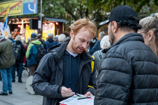 Das Bild zeigt einen Mann Mitte 40, Hannes Rockenbauch, der sich mit einem jungen Mann und einer jungen Frau auf dem Schlossplatz unterhält, um sie herum viele andere Menschen. Rockenbauch hat kurze rote Haare und einen kurzen roten Vollbart, er trägt eine schwarze Regenjacke, in seiner Hand hält er ein Klemmbrett mit Unterschriftenliste.