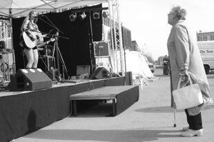 Alte Frau schaut auf eine Bühne, auf der eine junge Frau mit langen Haaren steht und Gitarre spielt