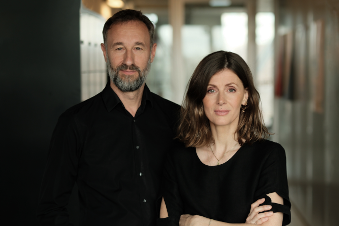 Andreas Marggraf links in schwarzem Hemd mit graumeliertem Bart und Aline Lüllmann rechts daneben mit vor dem Bauch verschränkten Armen, schulterlangen braunen haaren und einem schwarzen Oberteil, das knapp über den Ellenbogen endet und dort je ein eingenähte Ausparung hat. Im Hintergrund sieht man verschwommen Schließfächer.
