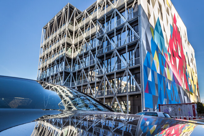 Foto des taz-Gebäudes aus Stahl und Glas mit buntem, geometrischen Kunstwerk an der Seitenwende. Im unteren Bildabschnitt spiegelt sich das Haus in der Motorhaube eines Autos.