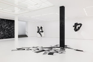 In einem mit weißem Papier ausgekleideten Ausstellungsraum verlaufen um eine tiefschwarze Säule breite Pinselstriche in verschiedenen Grau-und Schwarztönen auf dem Boden. Im hintergrund sind eine abstrakte Wandskulptur und ein gerahmtes Kohletuschegemälde zu sehen, das in einem schrägen Winkel von der Decke hängt und nur minimal den Boden berührt.