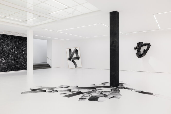 In einem mit weißem Papier ausgekleideten Ausstellungsraum verlaufen um eine tiefschwarze Säule breite Pinselstriche in verschiedenen Grau-und Schwarztönen auf dem Boden. Im hintergrund sind eine abstrakte Wandskulptur und ein gerahmtes Kohletuschegemälde