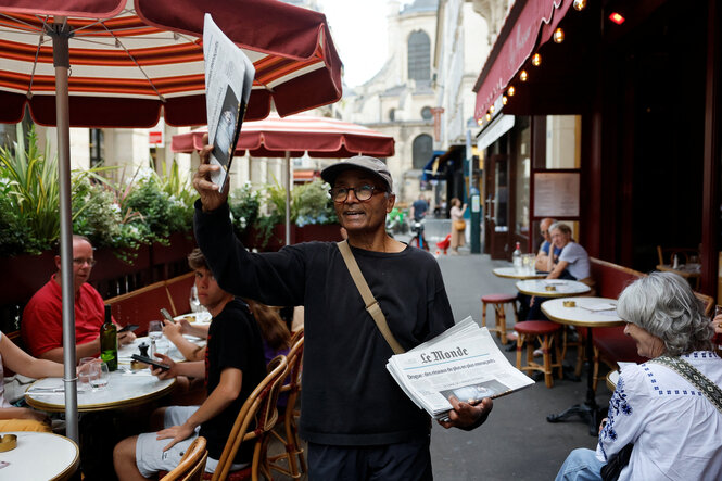 Eine Person steht auf einer Straßencafé-Terrasse und hält Zeitungen in der Hand. Im Hintergrund sitzen Menschen an Tischen unter roten Sonnenschirmen, die Straße ist von alten Gebäuden gesäumt.