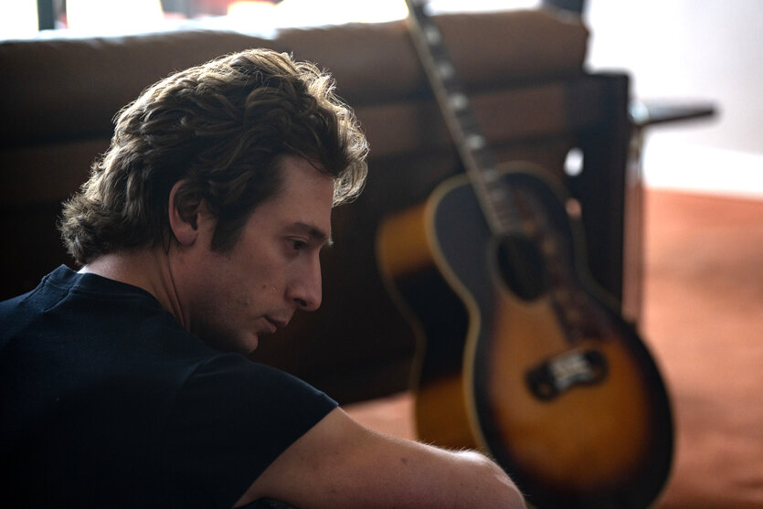 Der "Boss" (Jeremy Allen Walker) sitzt im Wohnzimmer auf dem Boden, im Hintergrund eine Gitarre.
