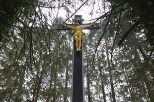 Mitten in einem Waldstück ragt ein hohes schwarzes Kreuz empor, darauf eine goldene Jesusfigur.