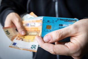 Nahaufnahme zweier Hände: In der einen eine blaue Gesundheitskarte, in der anderen Bargeldscheine.