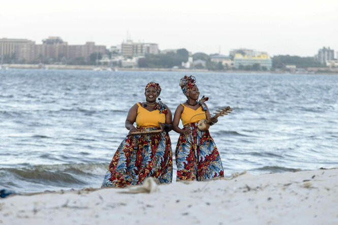 Pendo und Leah Zawose stehen in traditioneller Kleidung an einem Strand. Hinter ihnen ist das Wasser.