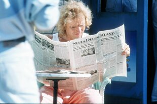 Der junge blonde Thomas Gottschalk sitzt in der Sonne auf einem Stuhl mit Bistrotisch und liest ganz konzentriert die Zeitung 