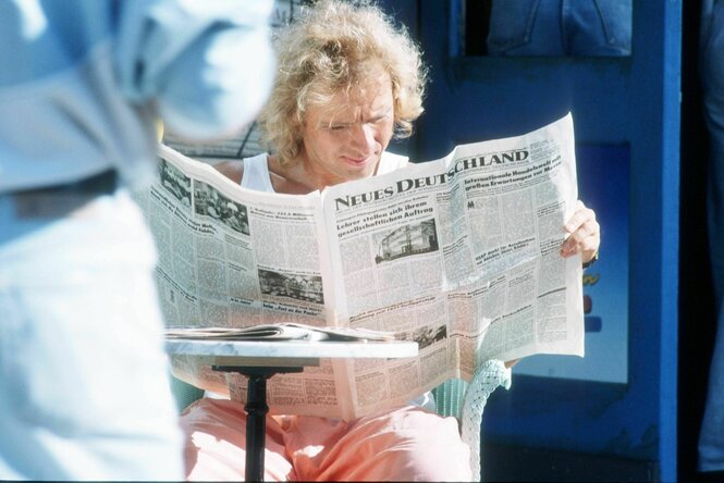 Der junge blonde Thomas Gottschalk sitzt in der Sonne auf einem Stuhl mit Bistrotisch und liest ganz konzentriert die Zeitung "Neues Deutschland"